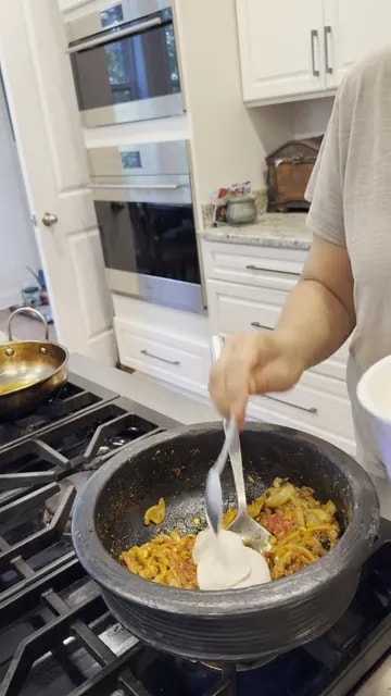Vegan yogurt being spooned into a spiced onion mixture in a black pan