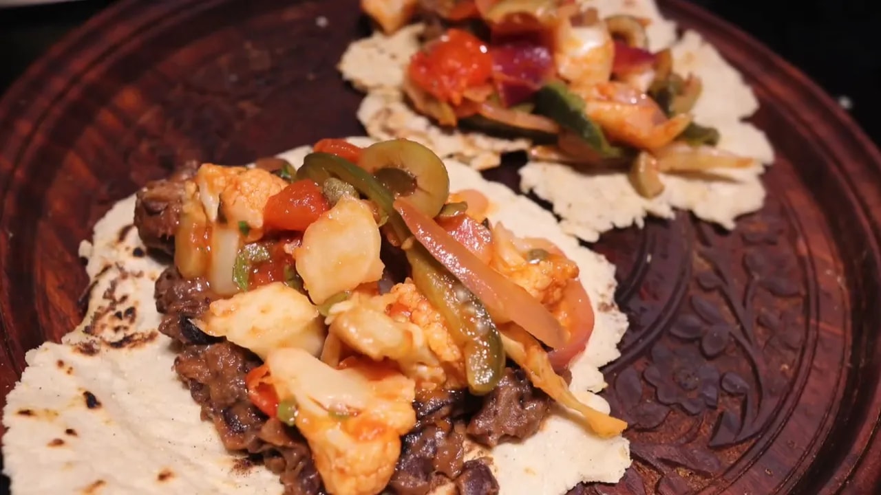Two corn tortillas topped with refried beans, cauliflower, tomatoes, onions, and peppers on a brown plate