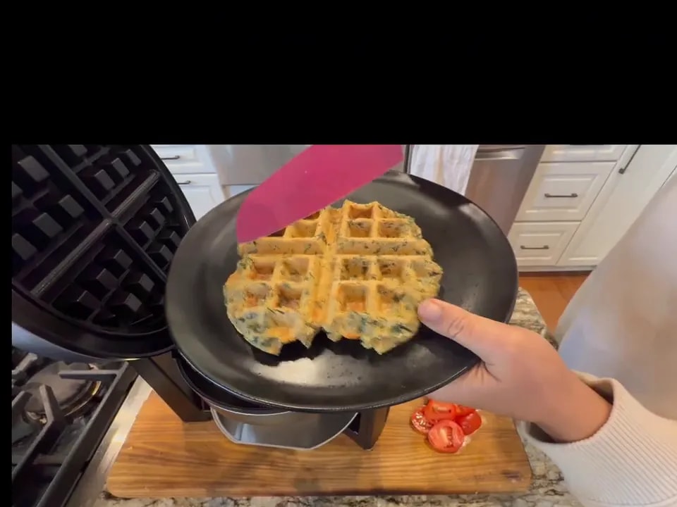 A golden spinach-flecked waffle on a black plate beside an open waffle maker