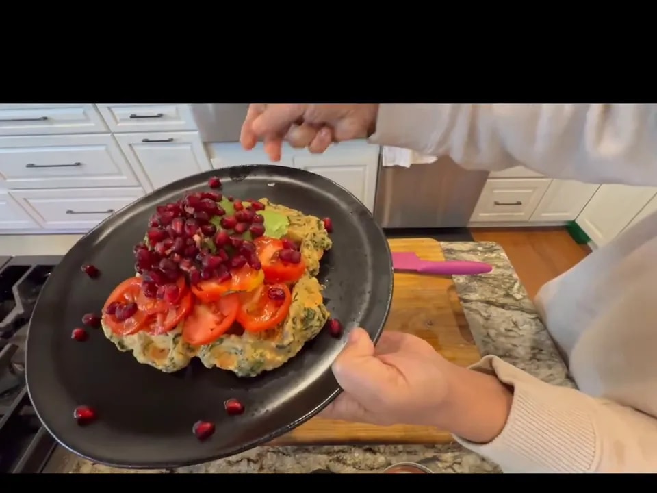 A lentil waffle topped with avocado, tomatoes, green chutney, and pomegranate seeds on a black plate