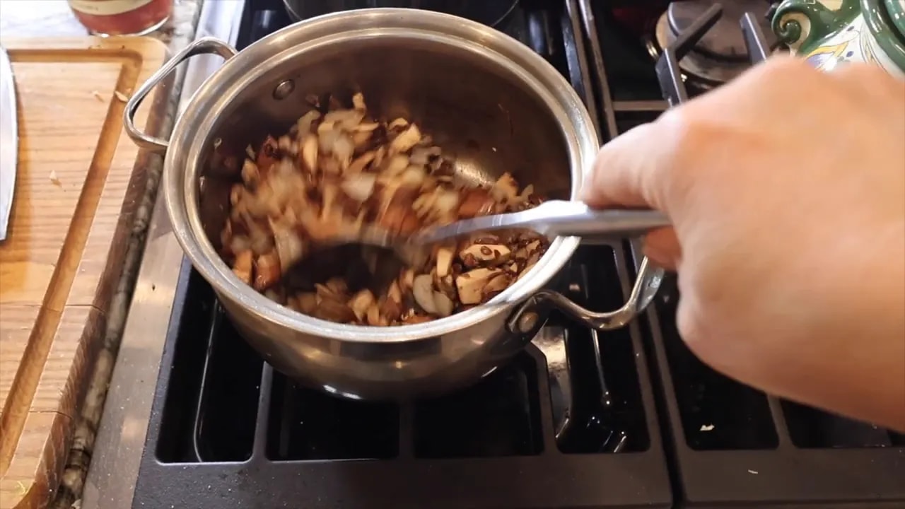 Diced mushrooms and onion in an uncovered stainless steel pot with a spoon resting inside